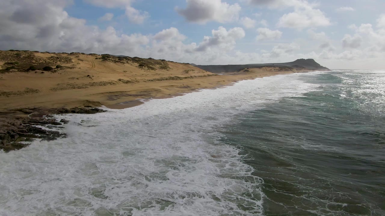 Foamy waves roll beside rocks toward a dune-backed sandy shoreline.