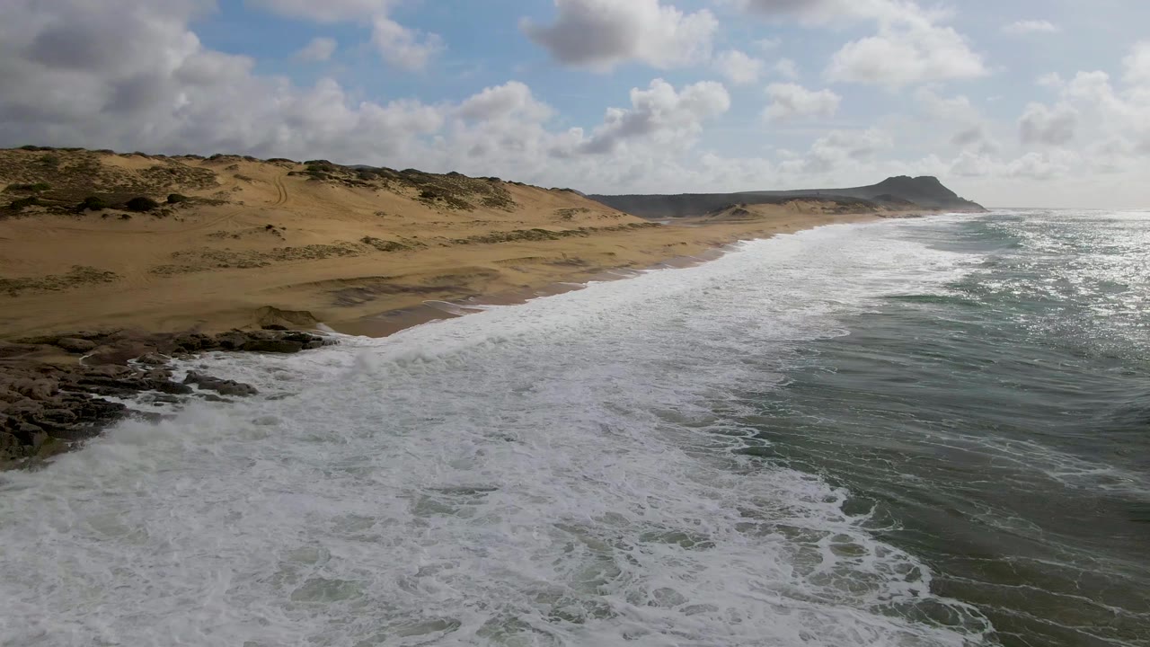At 0:02, whitewater fills more of the foreground near the rocks while the sandy Punta Gorda beach and dune ridge recede toward the distant headland, with no visible people.