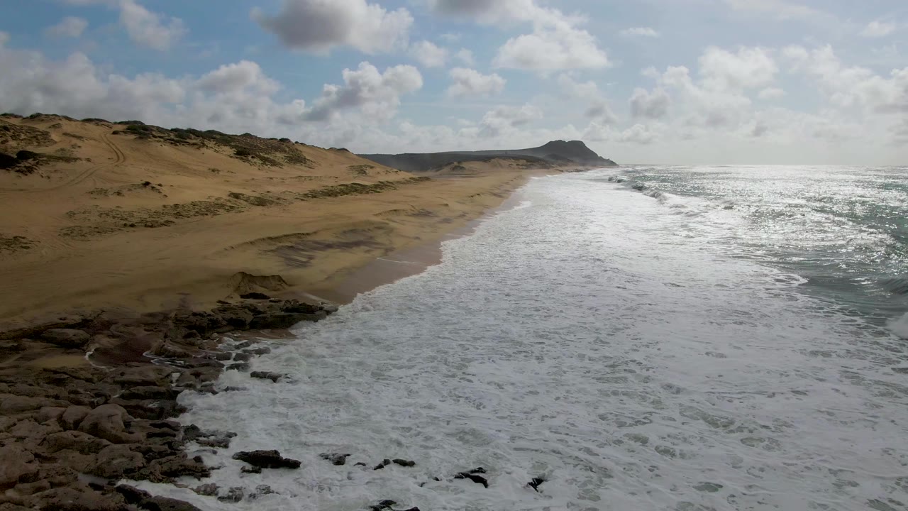 Aerial view over Punta Gorda, East Cape, with sandy dunes on the left, white surf along the beach, dark shoreline rocks below, and no visible people.