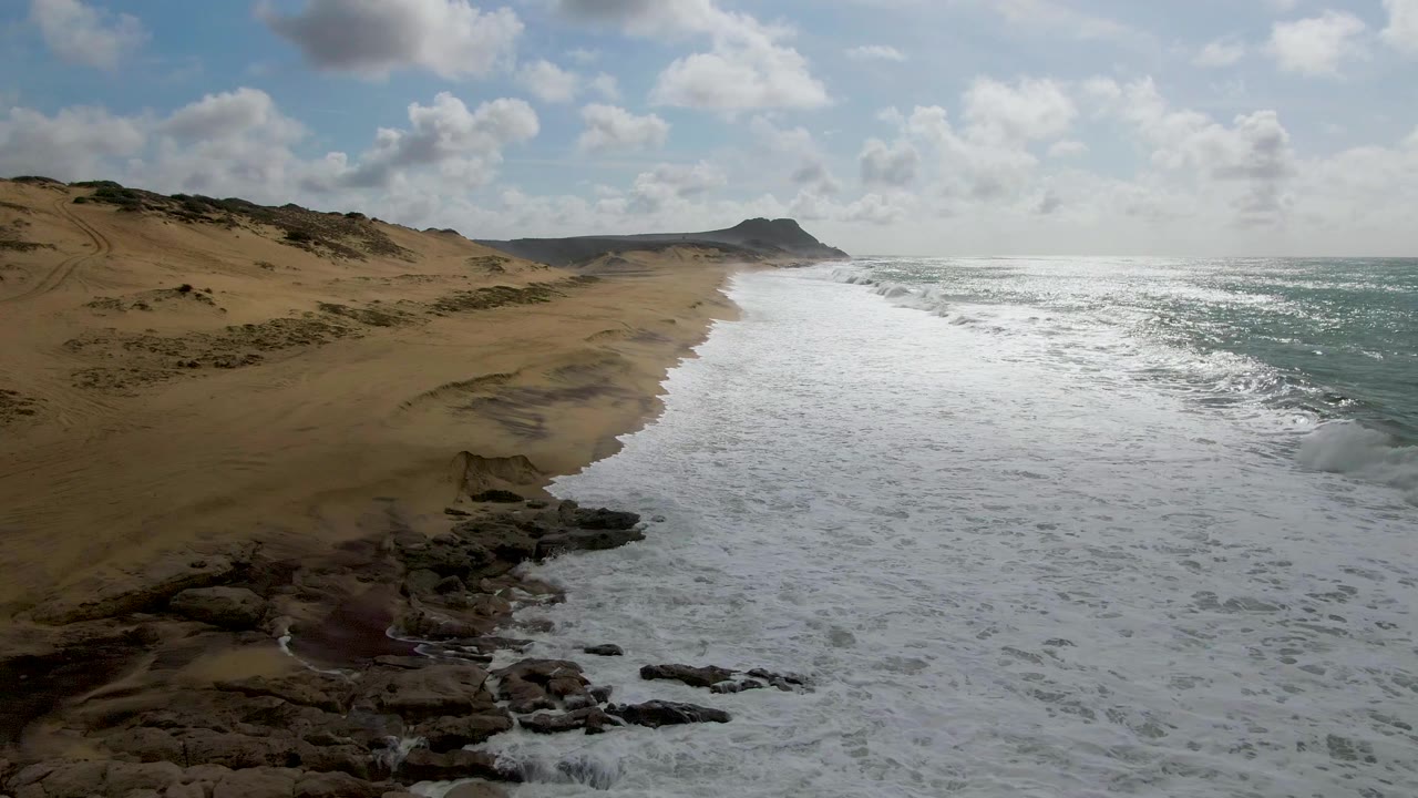 Wide aerial frame of the Punta Gorda beach with whitewater spread across the shore, exposed rocks near the lower center, dunes behind the sand, and no visible people.