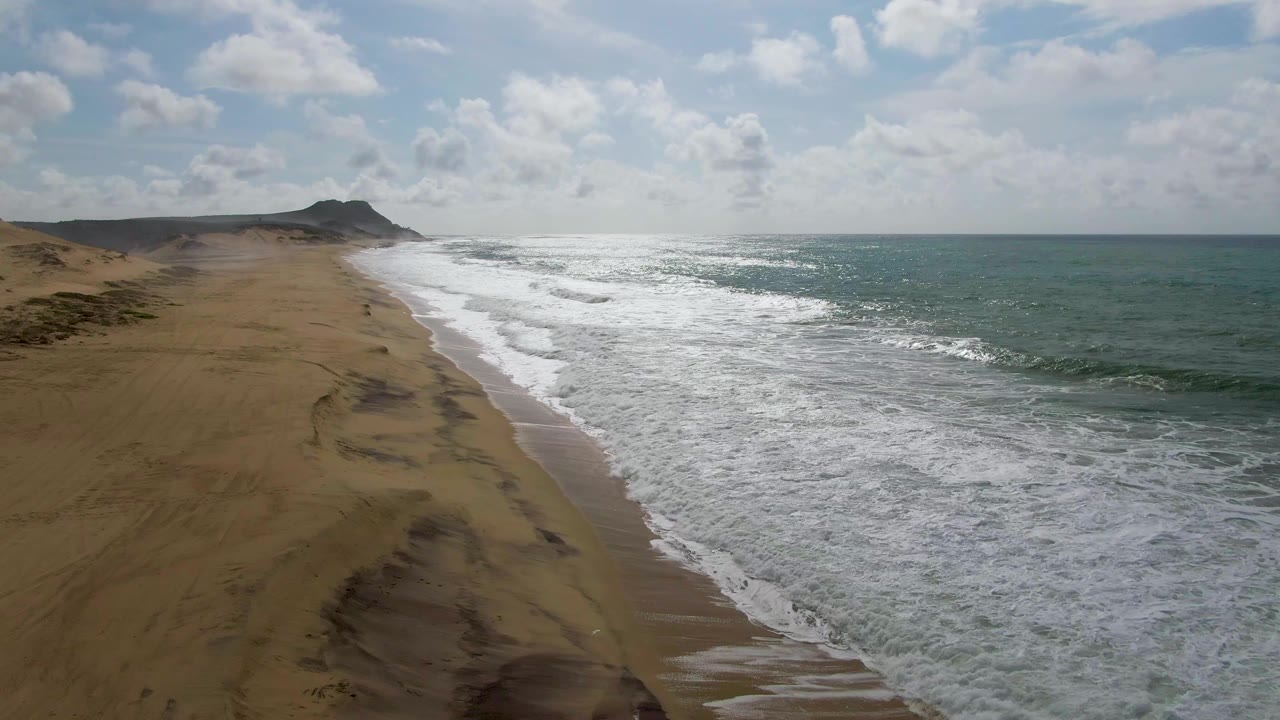 Opening aerial frame of Punta Gorda East Cape shoreline with sandy beach on the left, foamy surf in the center, green-blue ocean on the right, a distant headland, and no visible people.