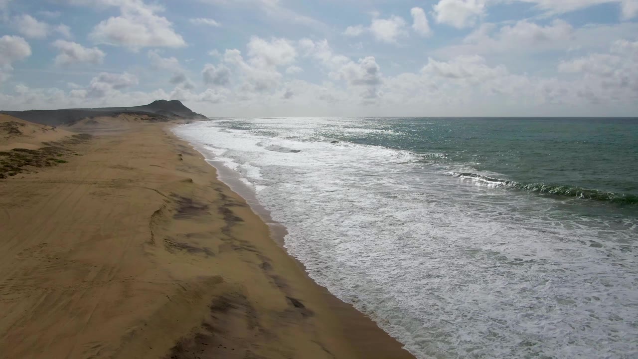 Foamy waves push farther up a long sandy shoreline with ocean to the right.
