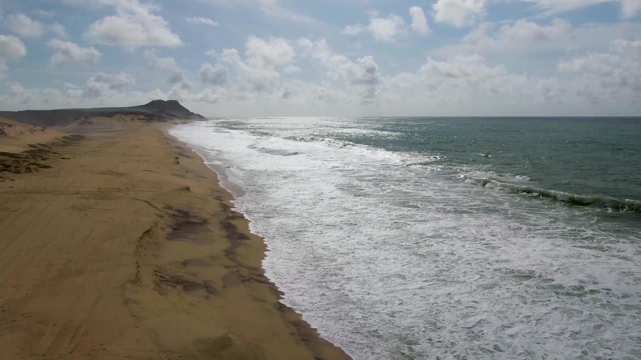 Aerial view over Punta Gorda East Cape with whitewater spreading across the middle of frame, empty brown sand on the left, darker ocean swells to the right, cloud cover overhead, and no visible people.