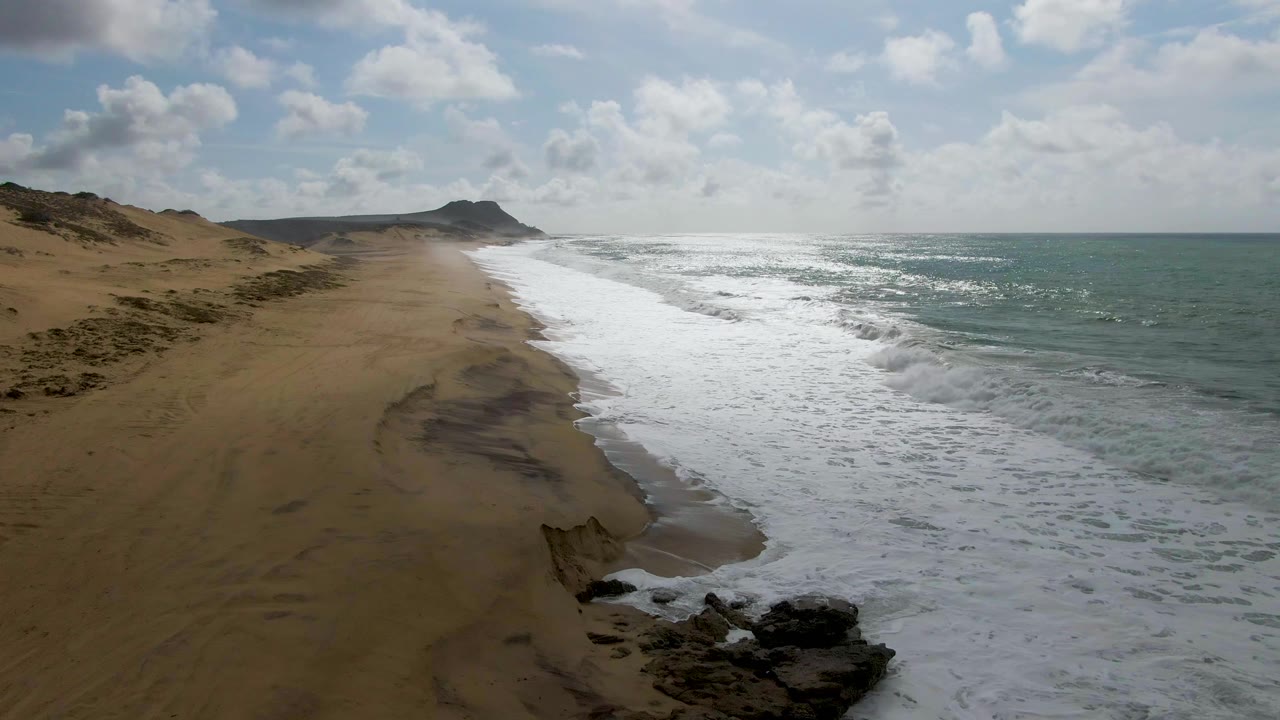 Aerial view of surf rolling beside a sandy beach and dunes.