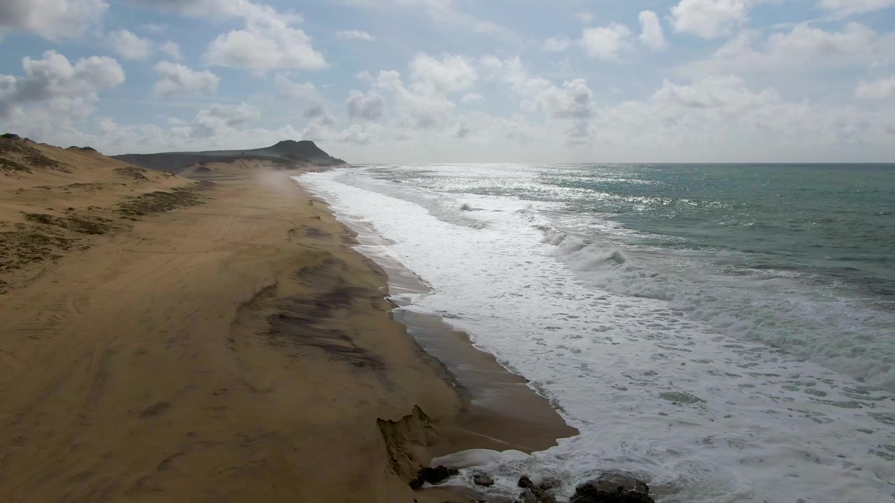 At 0:01 in Punta Gorda, East Cape, Baja California Sur, a broad band of white foam pushes up the beach while tan dunes and a distant hill remain on the left with no visible people.