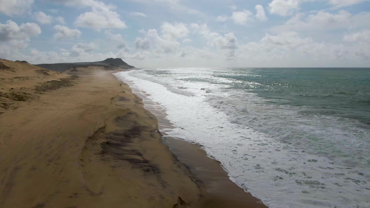At 0:02, the Punta Gorda shoreline runs diagonally through frame, with dunes and darker sand patches on the left, foamy surf in the center, ocean on the right, and no visible people.