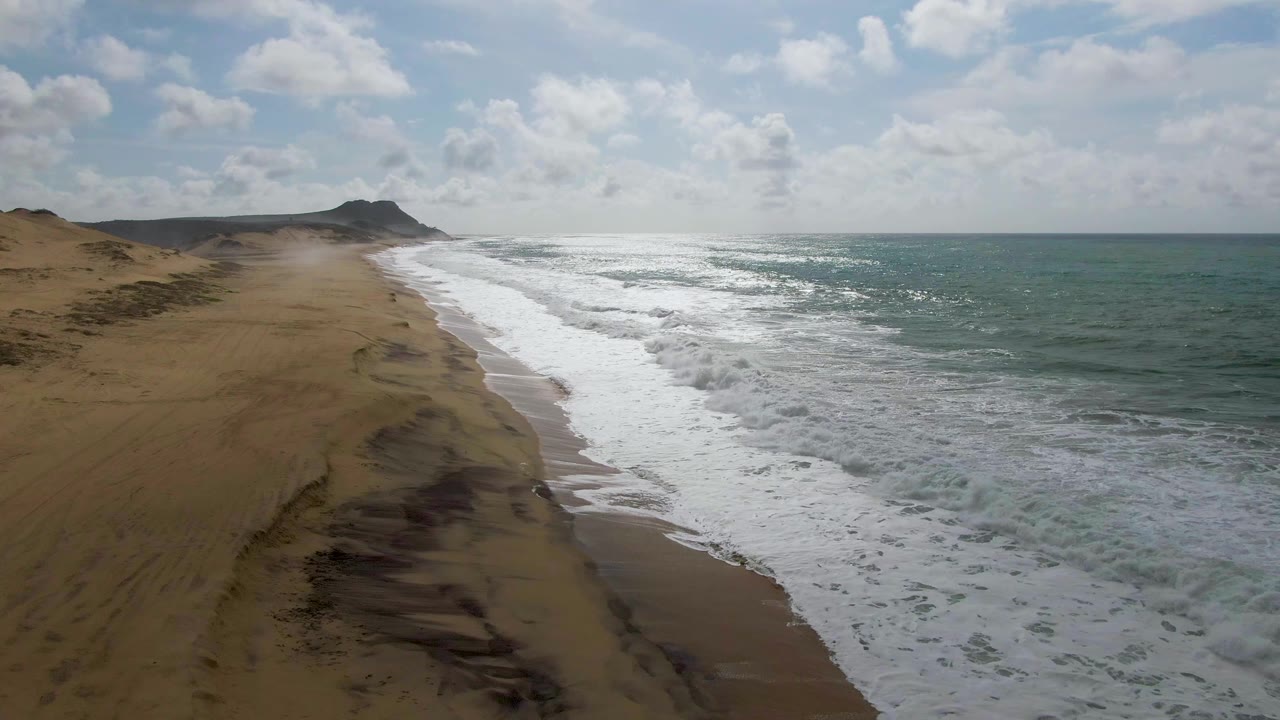 At 0:03 in Punta Gorda, East Cape, Baja California Sur, the aerial view shows an empty sandy beach, dunes to the left, choppy white water along the shore, blue-green ocean, and no visible people.