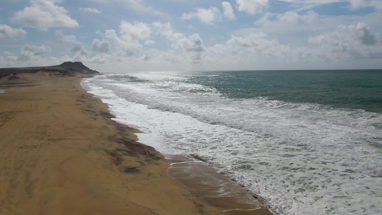 Aerial view of waves washing onto a sandy beach under a partly cloudy sky.