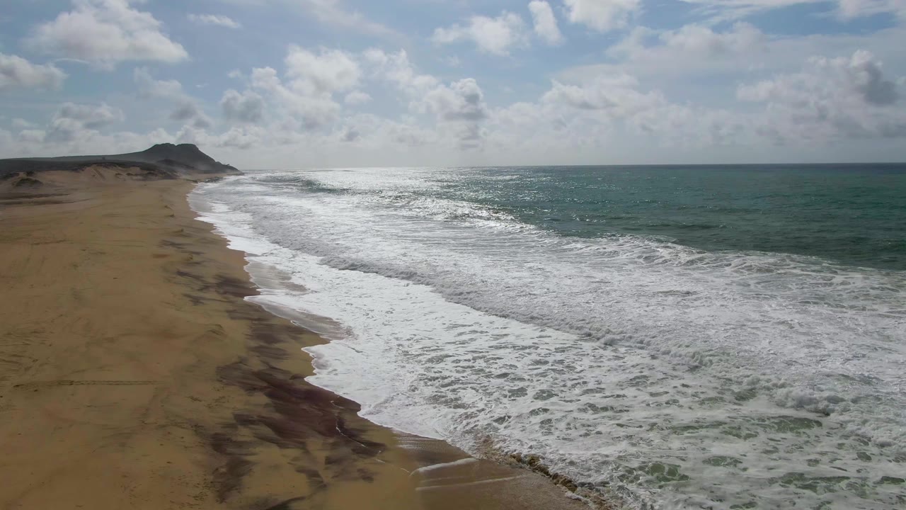At 0:01, the aerial view over Punta Gorda shows a wider band of white foam pushing onto the sand, with blue-green water on the right and no visible people on the beach.