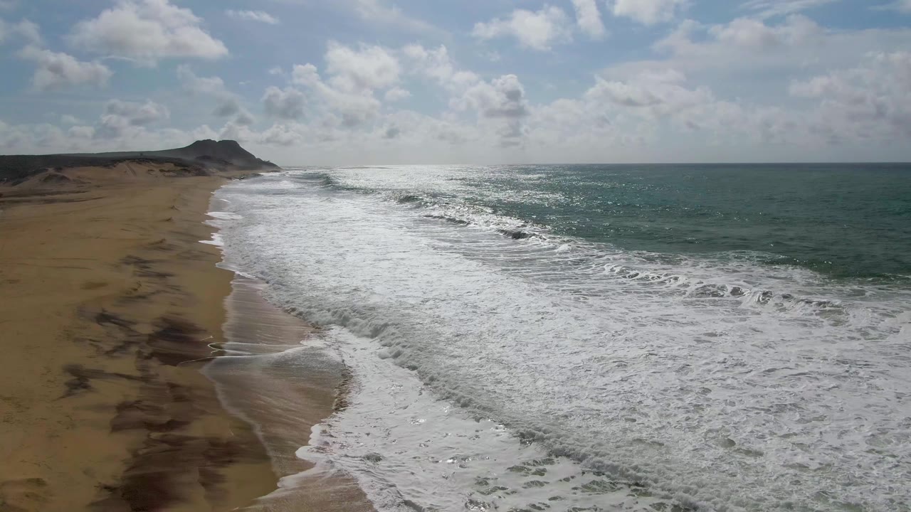 At 0:03, the aerial shot looks along the Punta Gorda beach as a bright sheet of foam covers the surf zone, with darker wet sand patches on the left and no visible people.