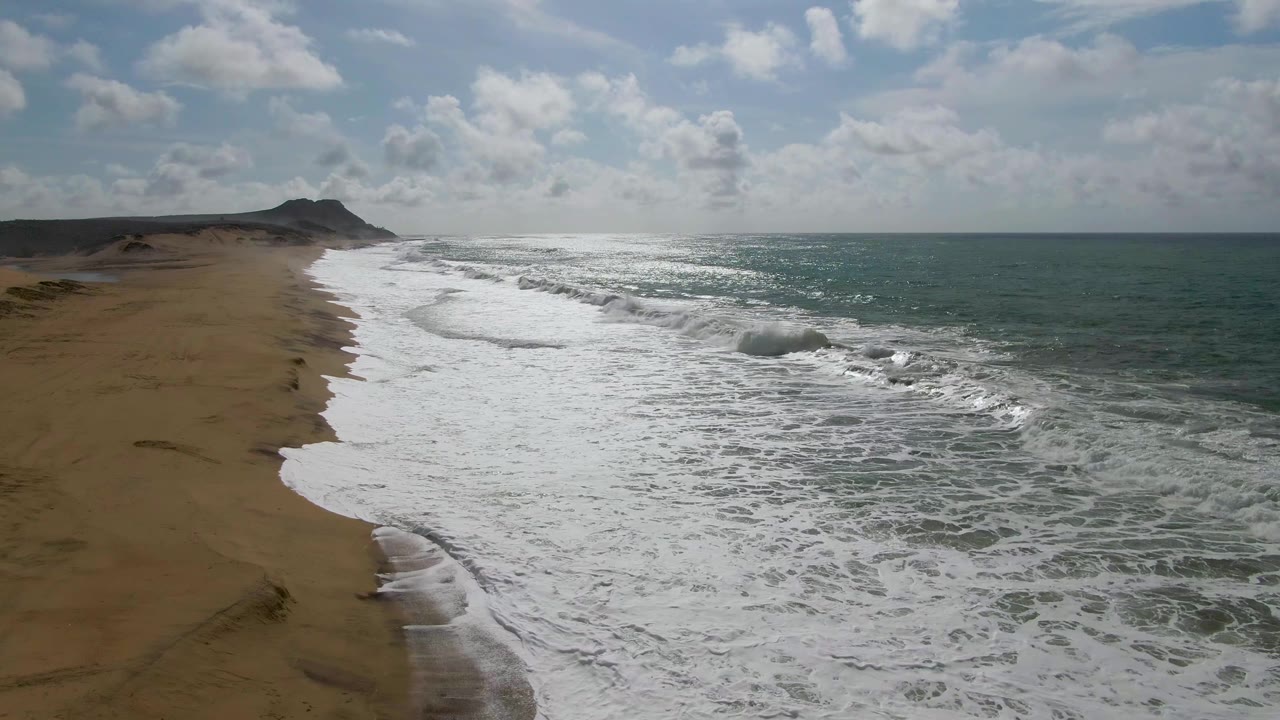 Aerial view of waves breaking along a sandy beach under scattered clouds.