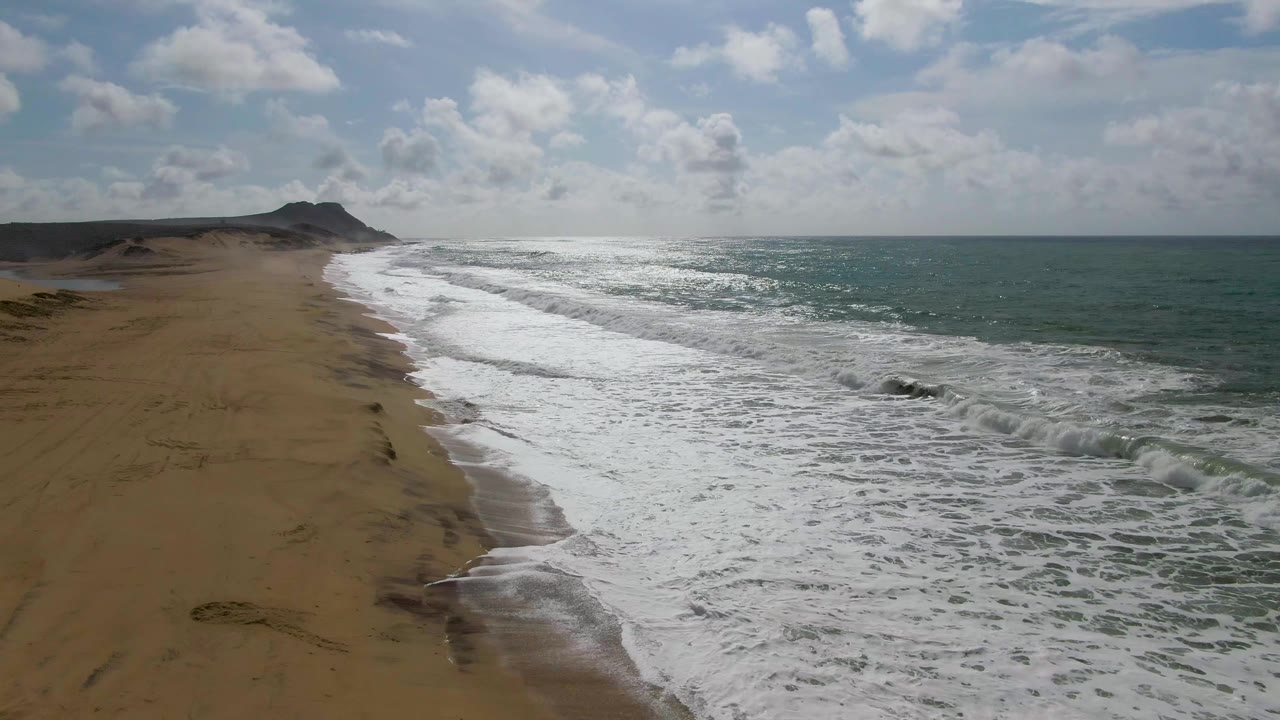 Drone-height view of Punta Gorda beach as diagonal wave sets break toward the sand, leaving white foam below a partly cloudy sky, with no visible people in the frame.