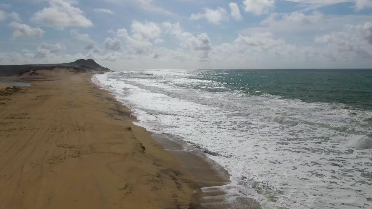 Aerial view along Punta Gorda's sandy coast with the beach filling more of the lower frame, white surf bands in the center, open ocean beyond, and no visible people.