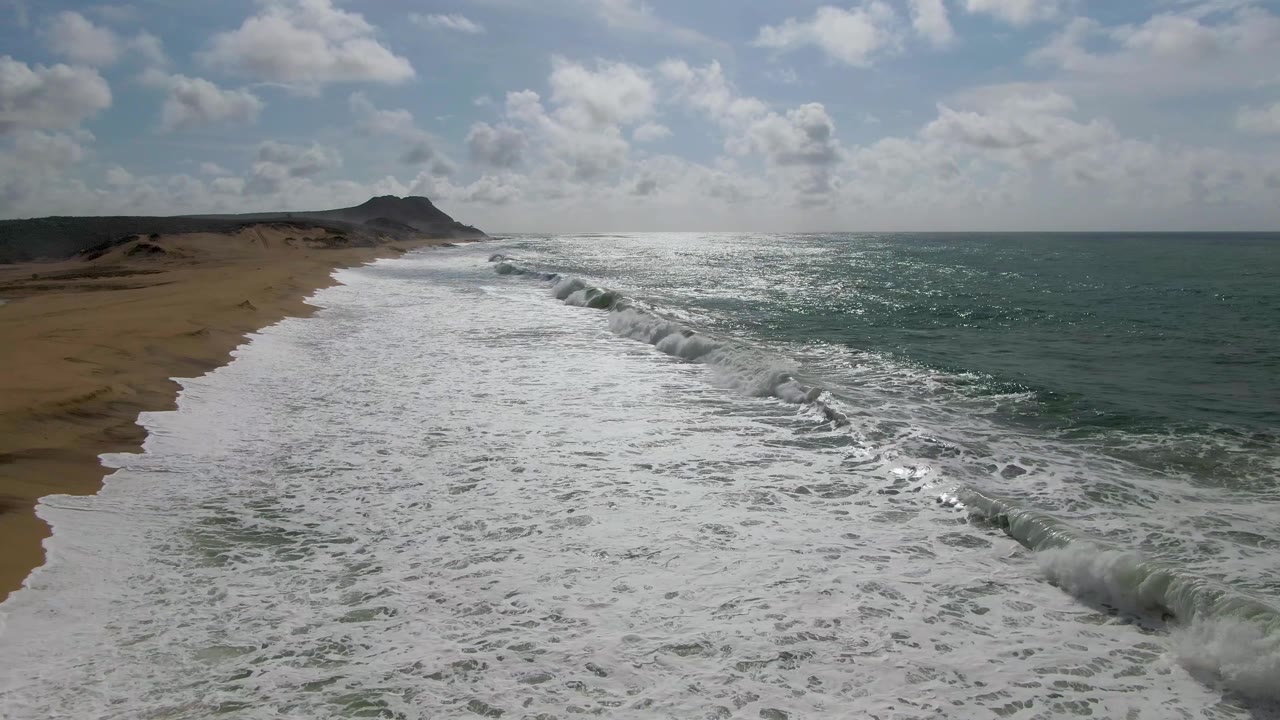 Aerial view over Punta Gorda, Baja California Sur, with white surf pushing onto the sandy beach, green-blue water to the right, low coastal hills in the distance, and no visible people.