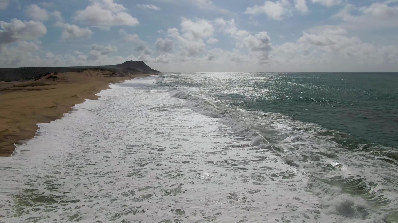 At 0:01, the Punta Gorda shoreline remains in an elevated diagonal view as a broader sheet of white foam covers the foreground beach and the sun glints across the ocean, with no visible people.