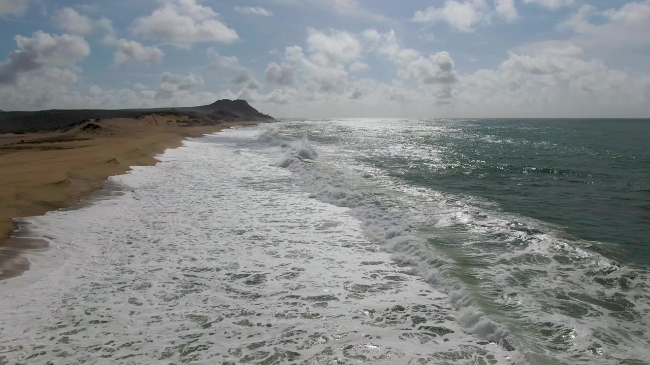 At 0:02, a fresh line of surf forms on the right side of the Punta Gorda beach while older foam spreads across the foreground sand, backed by low hills and a partly cloudy sky, with no visible people.