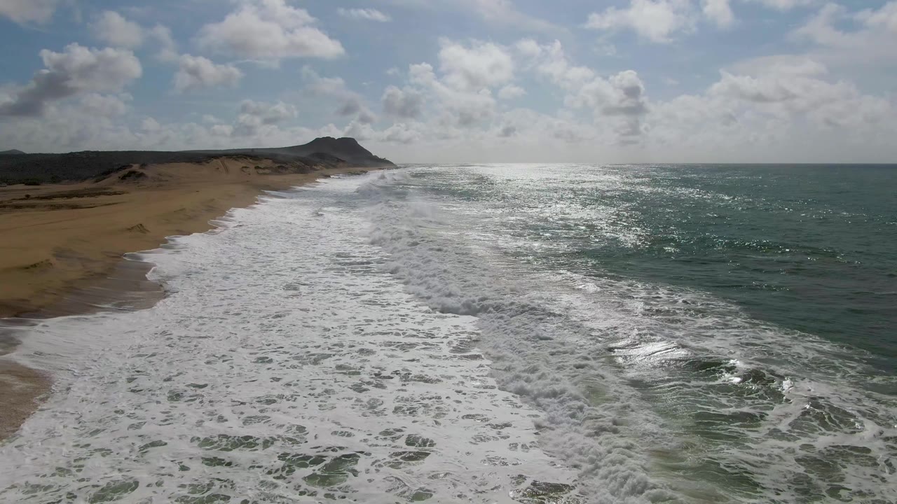 At 0:03, whitewater stretches down the empty Punta Gorda beach, with a darker green ocean channel on the right, sand and dunes to the left, and no visible people.