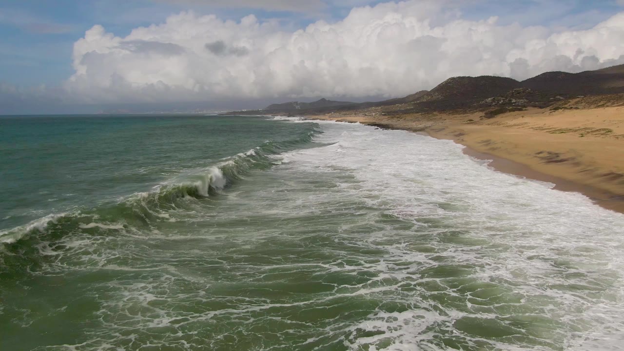 Aerial view of waves rolling toward a sandy beach under large clouds