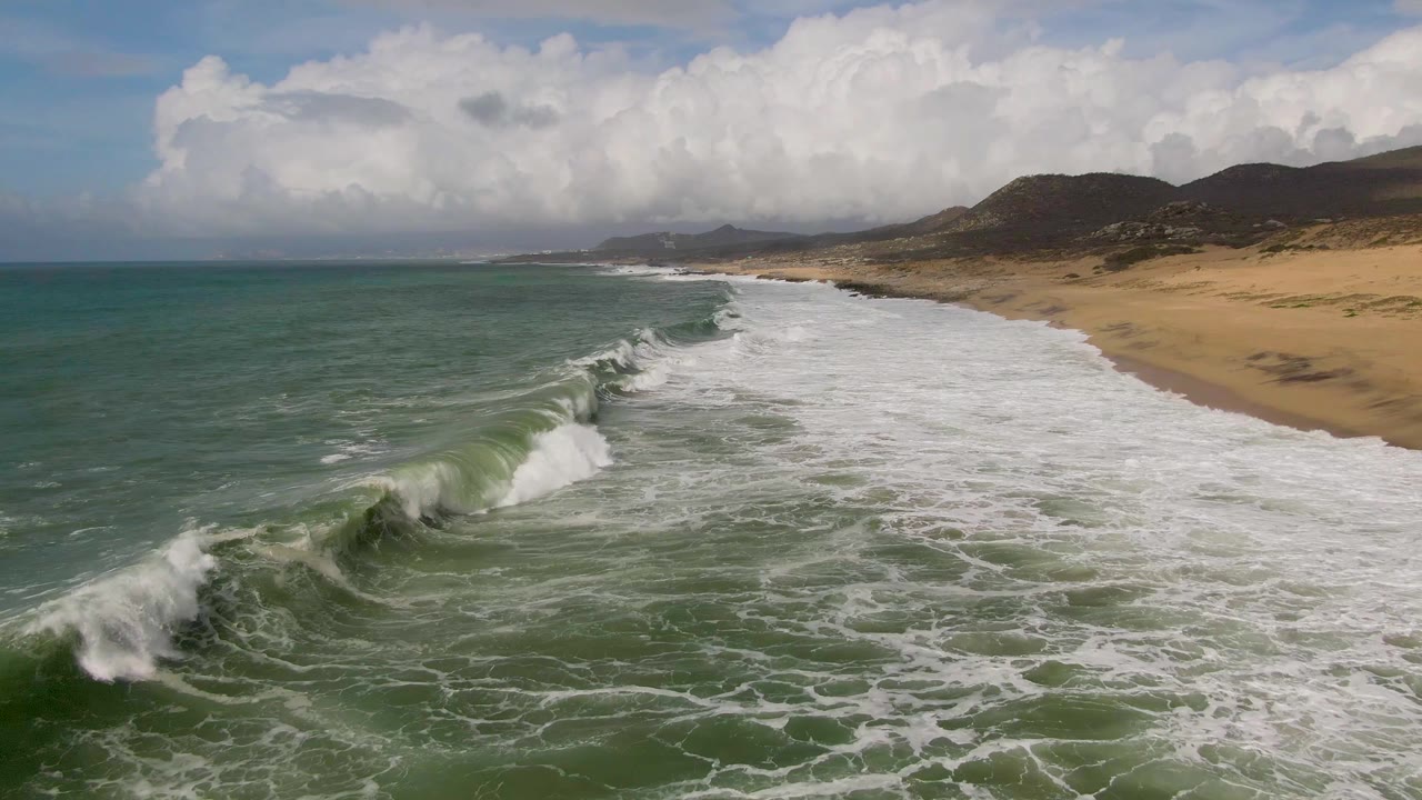 A wave crest lifts in the foreground at Punta Gorda, Baja California Sur, while broad white surf runs along the tan beach below distant hills, with no visible people.