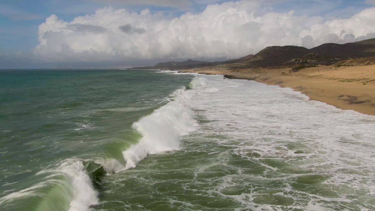 At Punta Gorda, Baja California Sur, a foreground wave breaks with white spray over green water, with a long sandy beach, dark hills, cloud cover, and no visible people.