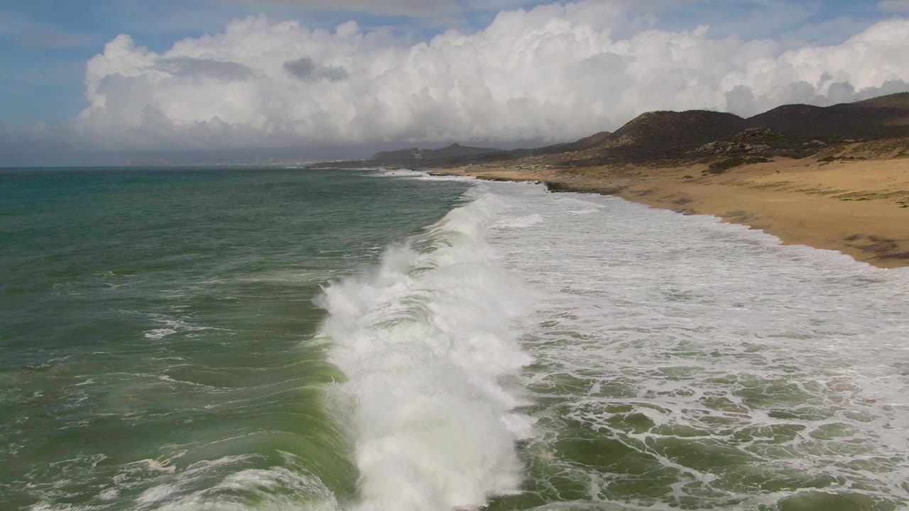 A large band of whitewater breaks diagonally across the foreground at Punta Gorda, Baja California Sur, with the empty sandy beach and coastal hills stretching into the background.