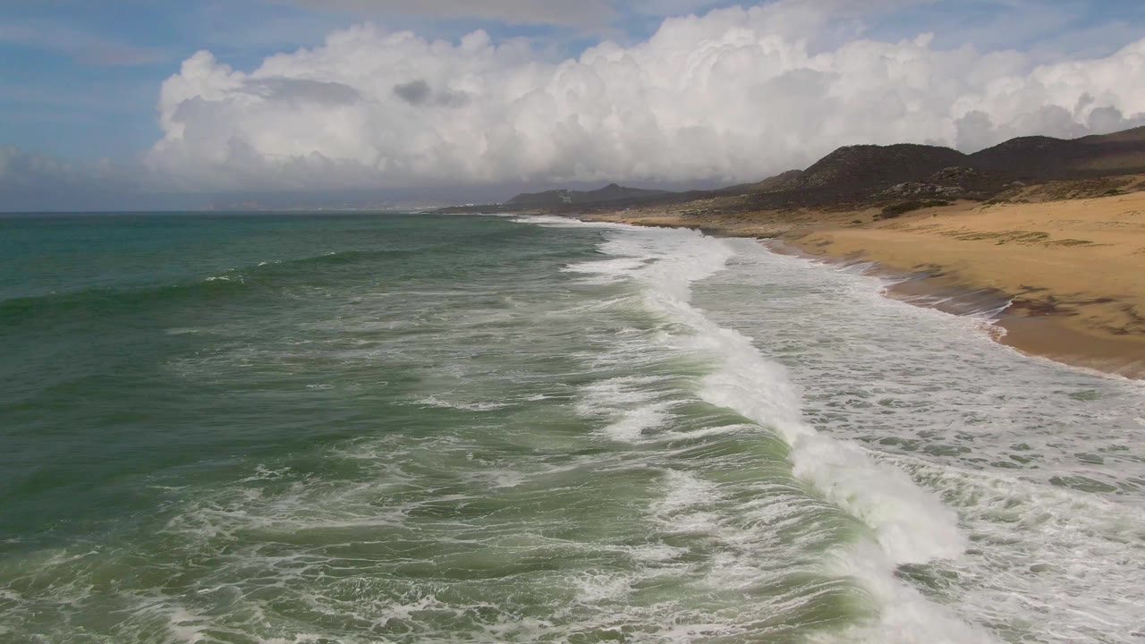 Aerial view at Punta Gorda, East Cape, Baja California Sur, with green-blue waves pushing white foam toward a sandy beach and low hills in the distance, no visible people.