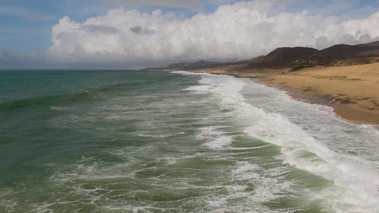 White surf spreads across green water beside a sandy shoreline.