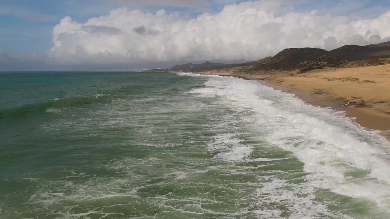 Aerial view of Punta Gorda surf with a broad white breaker crossing the midframe, open ocean on the left, sandy beach on the right, and no visible people.