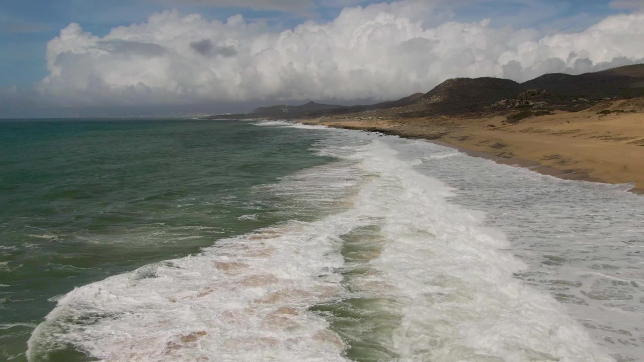 Opening aerial view of Punta Gorda, Baja California Sur, with green-blue ocean, white surf, tan beach, low hills, large clouds, and no visible people.