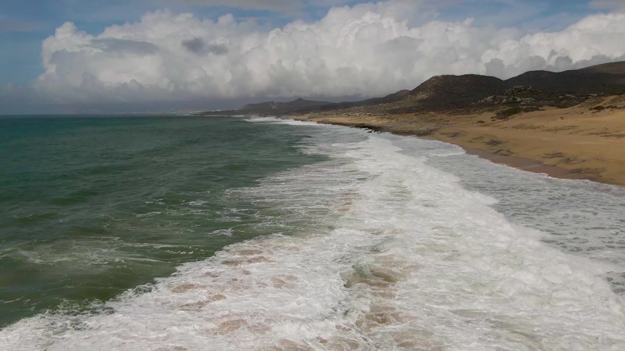Early frame from Punta Gorda shows a wider band of white foam pushing along the sandy beach, with low hills under bright cloud cover and no visible people.