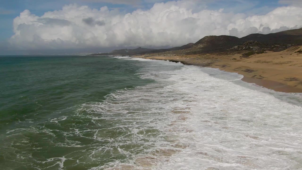 Long coastal view with ocean surf, sandy beach, hills, and clouds.
