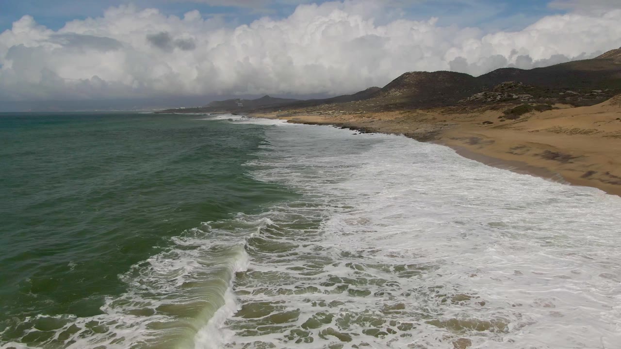 Aerial frame over Punta Gorda, Baja California Sur, with green ocean water pushing white foam onto a sandy shoreline, hills and clouds behind, and no visible people.
