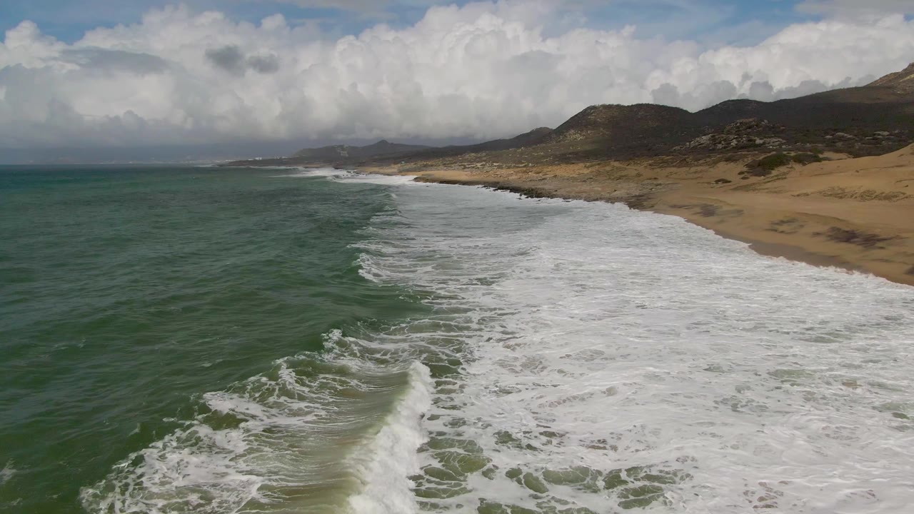 A second aerial moment at Punta Gorda shows more white foam crossing the sand as darker green water lines the left side and cloud-covered hills sit in the distance, with no visible people.