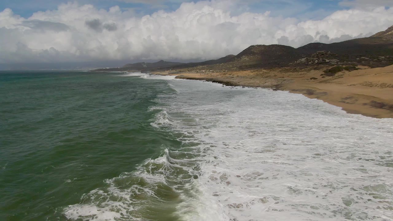 Aerial view of Punta Gorda coastline with a diagonal band of white surf, green water at left, tan beach at right, distant hills, heavy clouds, and no visible people.