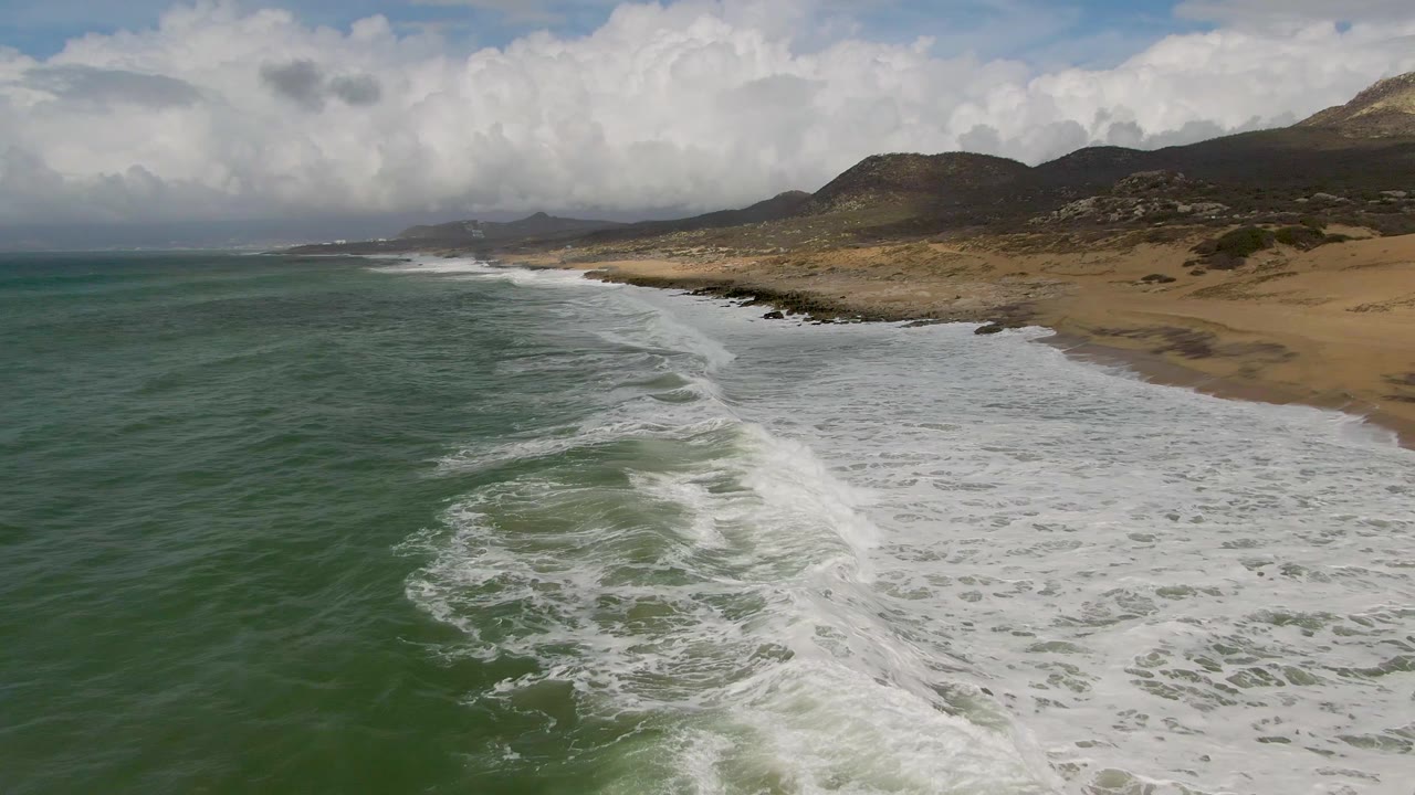 Aerial view of Punta Gordo East Cape surf rolling toward a sandy beach, with green water in the foreground, low hills in the distance, cloud cover overhead, and no visible people.