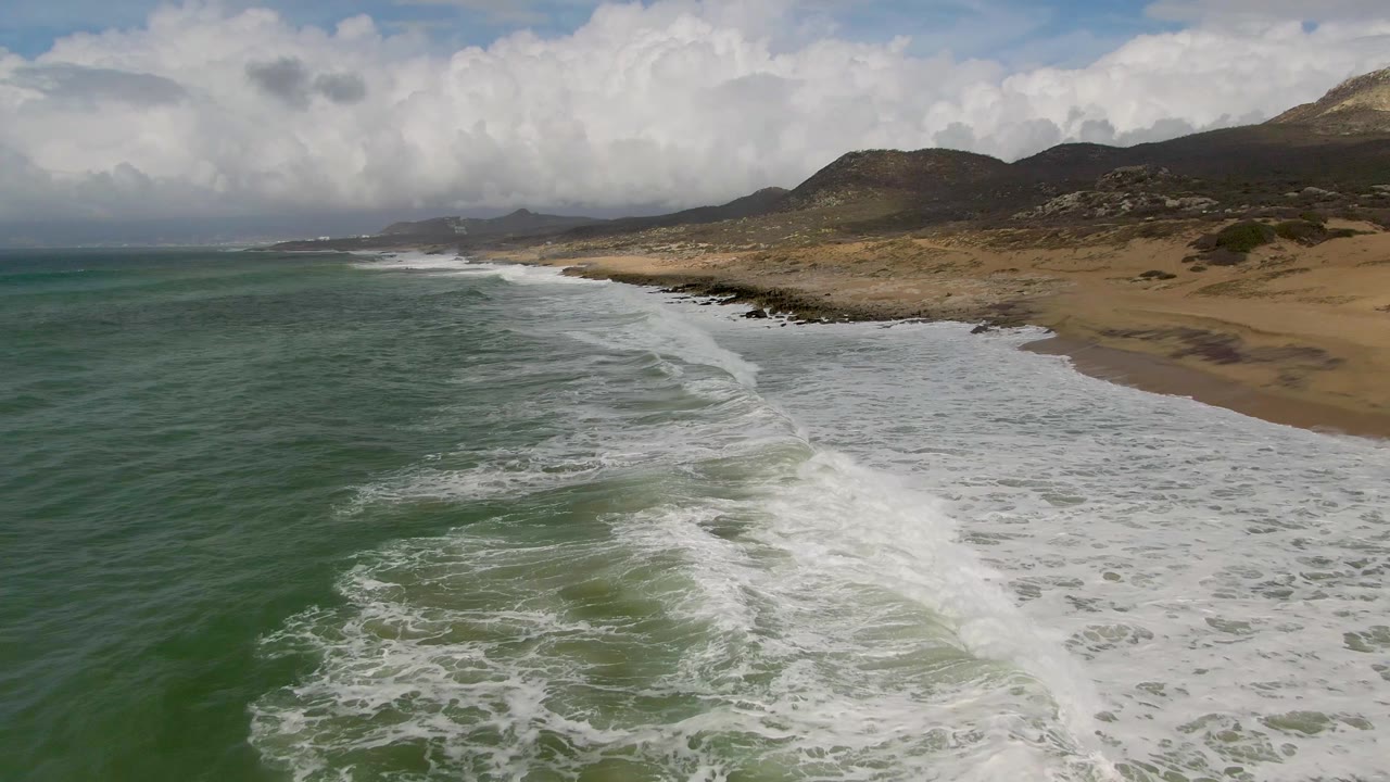 Punta Gordo East Cape shoreline with a broad band of white foam pushing farther onto the sand, green ocean to the left, low hills behind the beach, and no visible people.