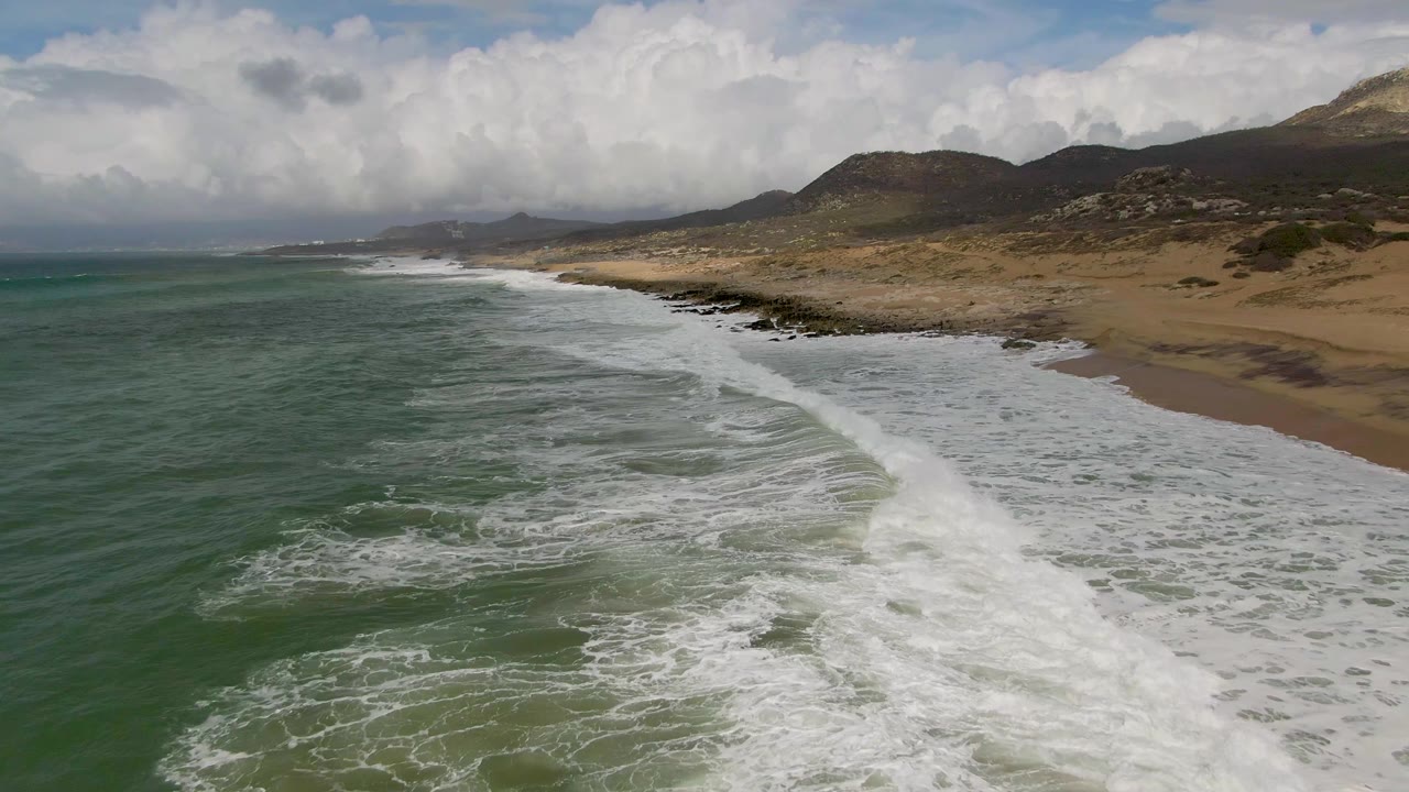 Aerial Punta Gordo East Cape coast view with diagonal white breakers, green water occupying the left side, sand and scattered shoreline rocks on the right, distant hills, and no visible people.