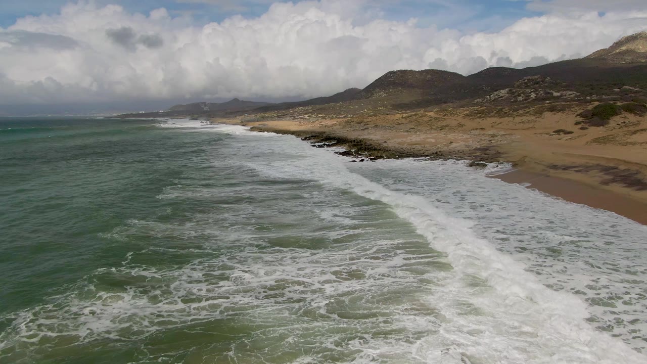 Wide aerial coastline with foamy surf, sandy beach, rocks, and hills beneath clouds.