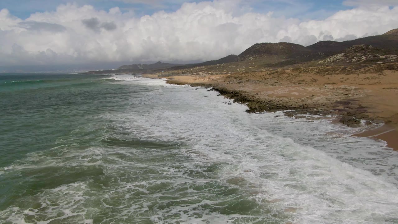Aerial view of green waves breaking along a rocky beach with hills and clouds beyond.