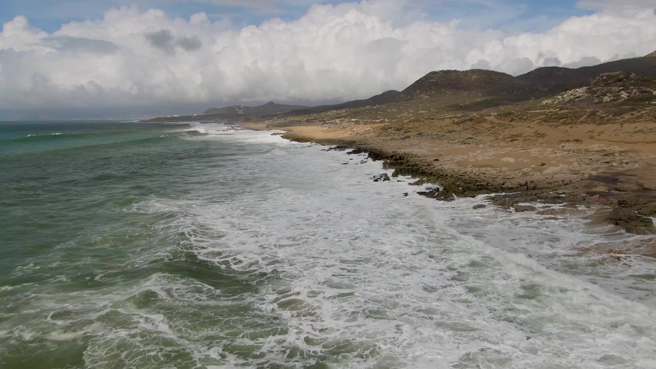 At 0:01, a broad band of white surf pushes farther up the Punta Gorda rocky beach, with green water to the left, brown hills to the right, and no visible people.