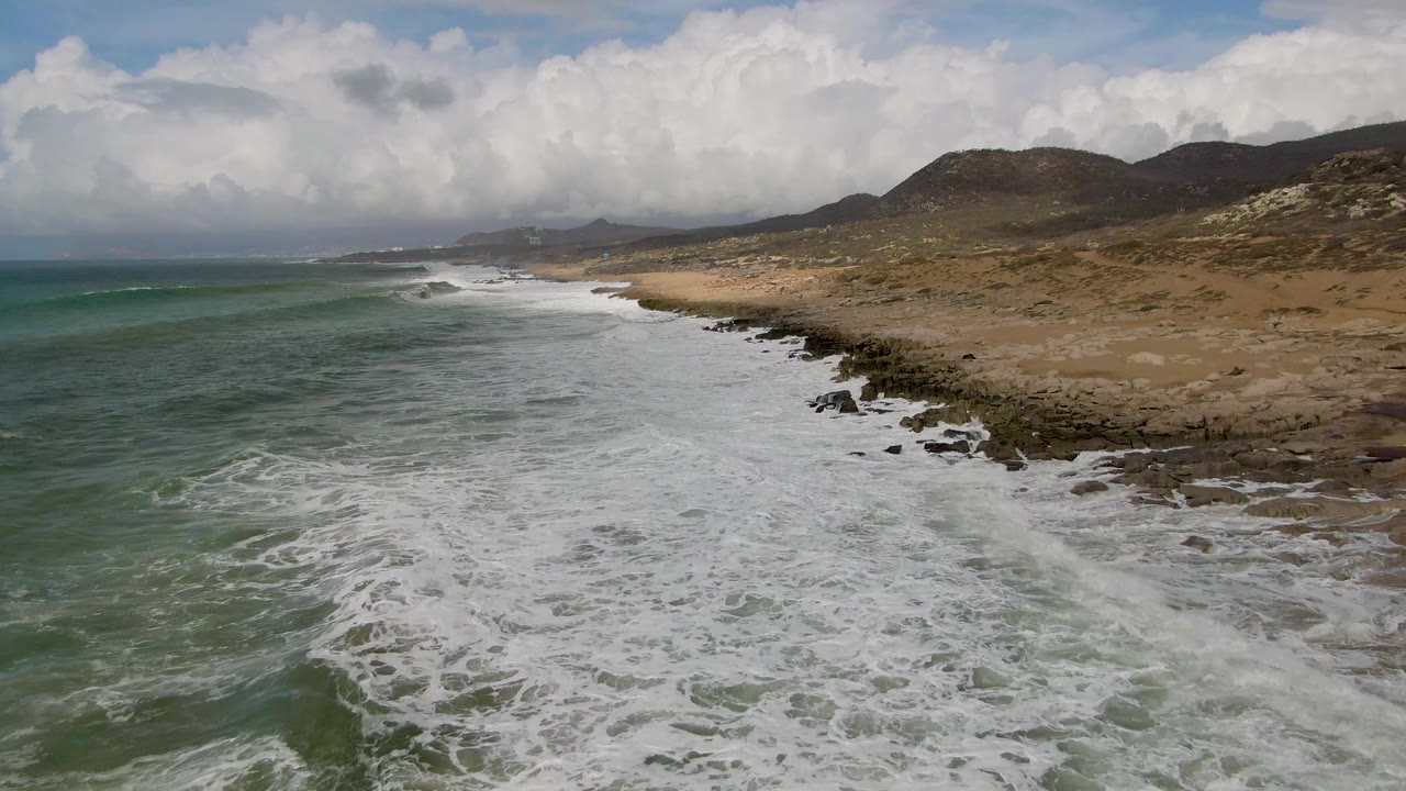 At 0:02, foamy water fills more of the lower frame while the Punta Gorda coast curves toward distant headlands under bright cloud cover, with no visible people.