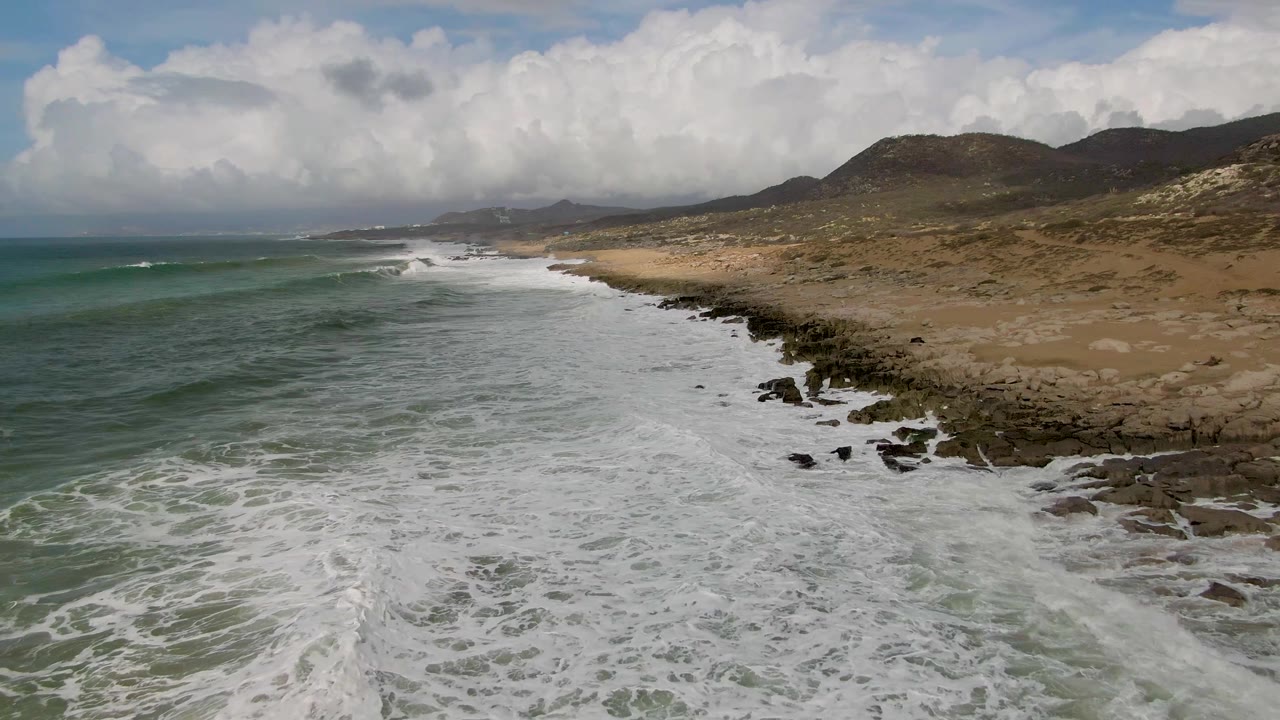 At 0:03, breaking waves form a long white strip beside the rocky Punta Gorda shore, with open water left, sand and scrubby hills right, and no visible people.
