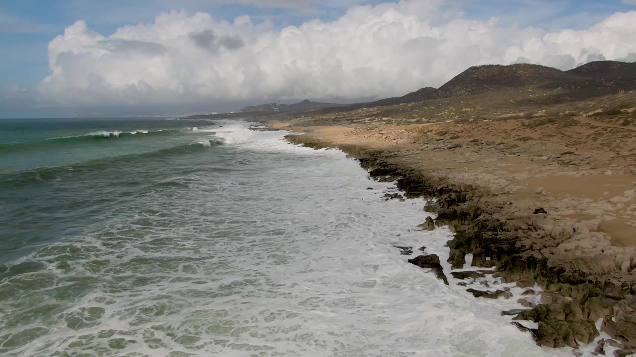 Aerial view of Punta Gorda East Cape surf spreading over a rocky and sandy shoreline, with low hills and large clouds in the background and no visible people.