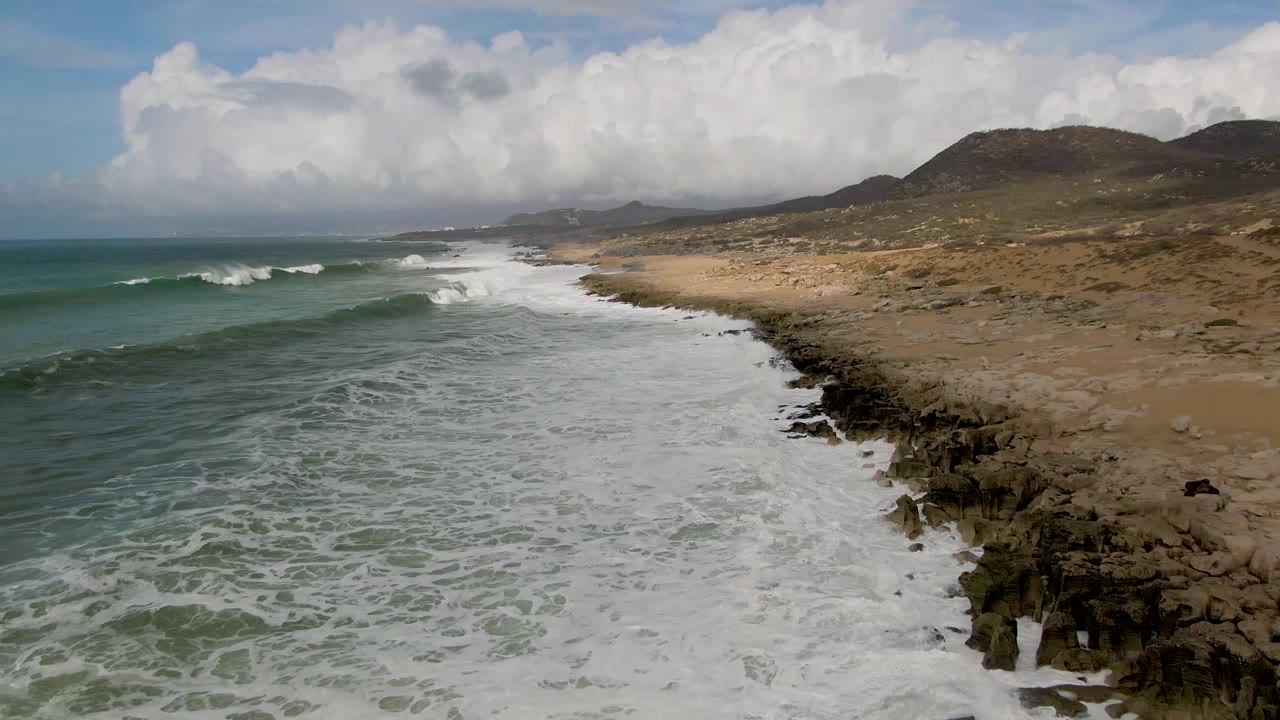 Punta Gorda East Cape waves roll in from the left and leave broad white foam across the nearshore water, with dark shoreline rocks, pale sand, distant hills, and no visible people.