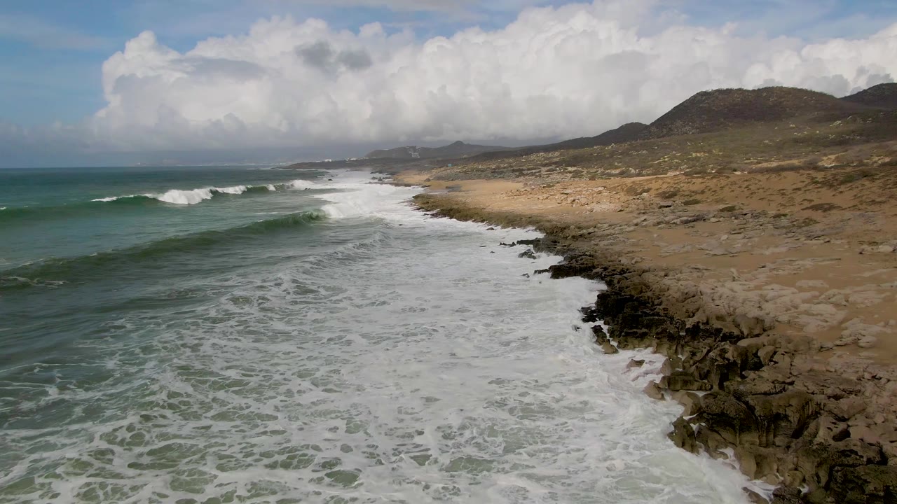 A breaking wave line at Punta Gorda East Cape advances along the shore, with foamy water below, dark rocks on the right, dry beach terrain, hills, and no visible people.