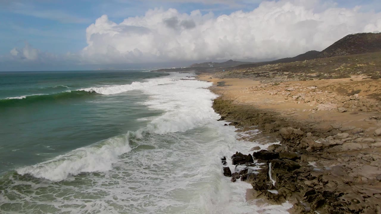 Punta Gorda East Cape surf rolls toward a sandy and rocky shoreline, with a broad white foam band, dark rocks near the lower right, distant coast, and no visible people.