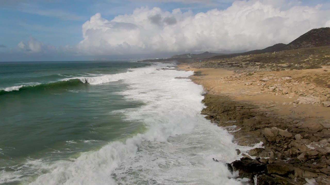 A wave crest breaks offshore at Punta Gorda East Cape while white wash spreads across the beach edge and rocky outcrops, with arid hills under a cloud-filled sky and no visible people.