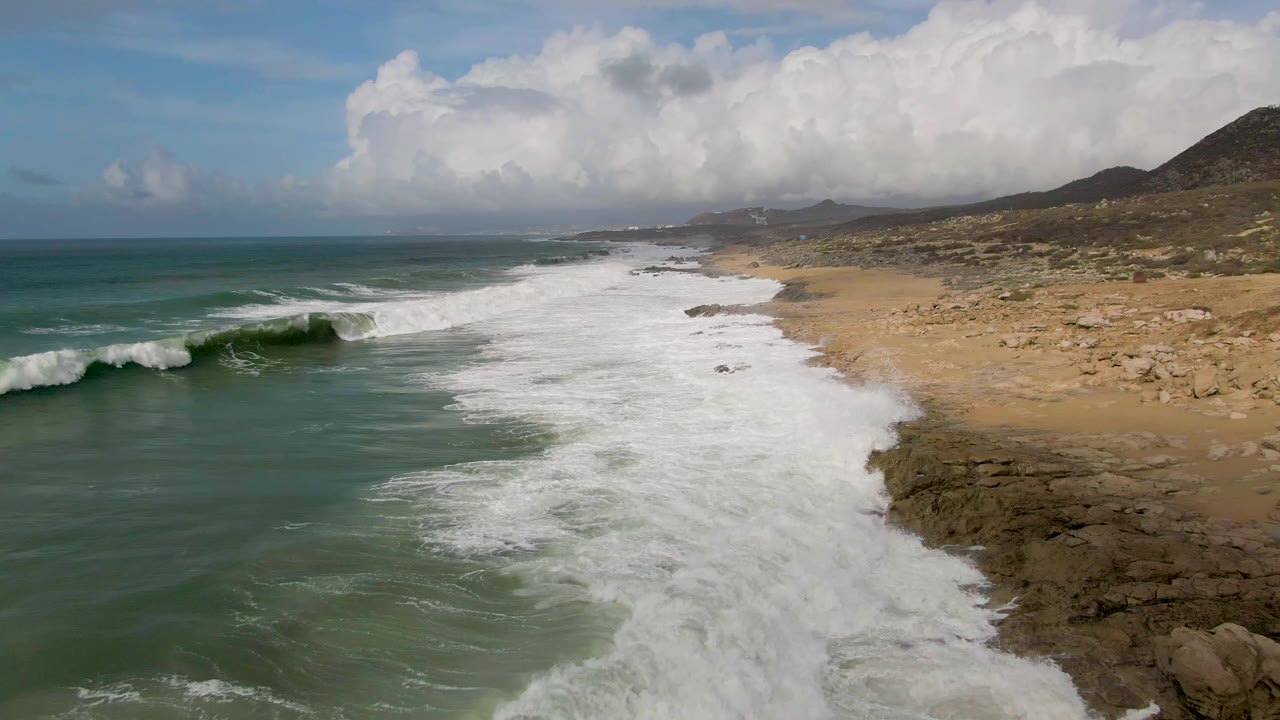 The Punta Gorda East Cape shoreline stretches diagonally through frame as waves approach from the left, white foam covers the beach margin, and no visible people appear on the sand or rocks.