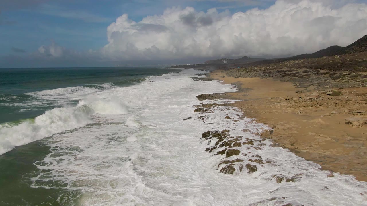 Aerial frame of Punta Gordo shoreline with white surf spreading across rocks and sand, teal ocean to the left, clouded hills in the distance, and no visible people.