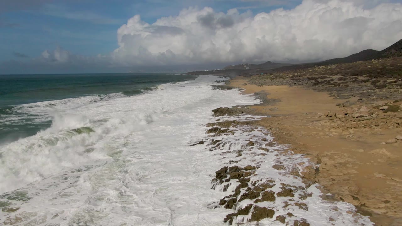 Aerial view in Punta Gordo as a larger band of whitewater pushes toward the rocky outcrops and sandy beach, with open ocean left and no visible people.